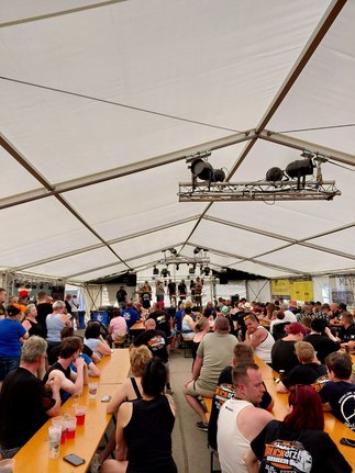 Menschen sitzen an langen Holztischen in einem großen Festzelt und schauen auf eine Bühne mit mehreren Personen
