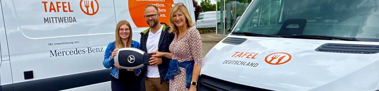Drei Personen stehen vor zwei weißen Lieferwagen mit dem Schriftzug 'TAFEL' und Mercedes-Benz-Logo, eine Person hält ein großes Lenkrad mit Mercedes-Stern