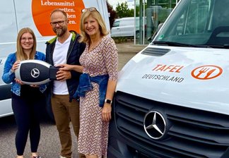 Drei Personen stehen vor zwei weißen Lieferwagen mit dem Schriftzug 'TAFEL' und Mercedes-Benz-Logo, eine Person hält ein großes Lenkrad mit Mercedes-Stern