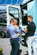 Drei Männer stehen vor einem weißen Mercedes-Benz Lkw mit geöffneter Fahrertür und unterhalten sich auf einem gepflasterten Platz.
