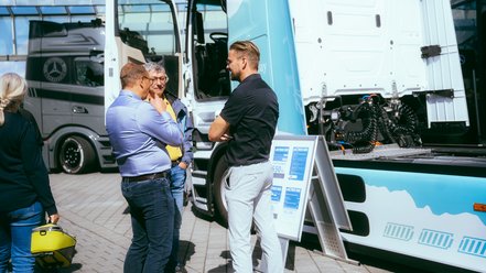 Drei Männer stehen vor einem weißen Mercedes-Benz Lkw mit geöffneter Fahrertür und unterhalten sich auf einem gepflasterten Platz.