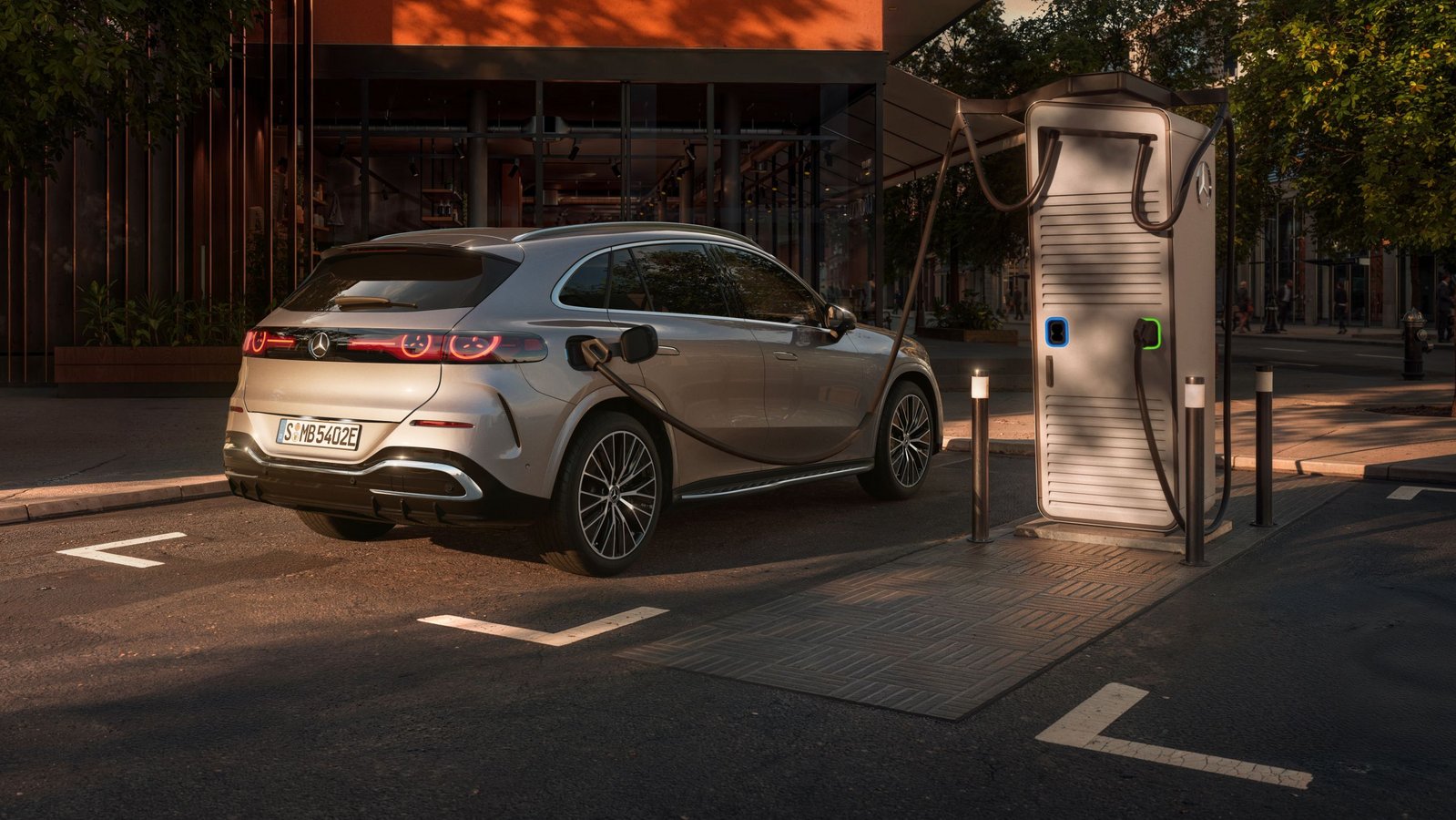 Silberner Mercedes-Benz GLC EQ SUV lädt an einer Ladestation vor modernem Gebäude mit vertikalen Holzlamellen bei Sonnenuntergang.
