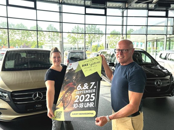 Zwei Personen halten ein Plakat für das Bike Festival 2025 am 6. und 7. September von 10 bis 18 Uhr im Kurort Oberwiesenthal, stehend zwischen zwei Mercedes-Benz Fahrzeugen in einem Autohaus.