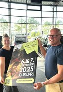 Zwei Personen halten ein Plakat für das Bike Festival 2025 am 6. und 7. September von 10 bis 18 Uhr im Kurort Oberwiesenthal, stehend zwischen zwei Mercedes-Benz Fahrzeugen in einem Autohaus.