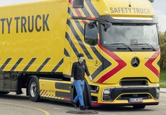 Gelber Mercedes-Benz Actros Lastwagen mit schwarzem und rotem Chevron-Muster und der Aufschrift 'SAFETY TRUCK', davor steht eine Person in blauer Hose und schwarzer Jacke auf einem Asphaltplatz.