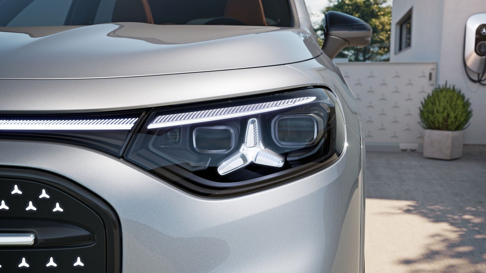 Nahaufnahme des vorderen linken Scheinwerfers und Teils des Kühlergrills eines silbernen Mercedes-Benz GLB EQ vor einem Haus mit Ladestation.