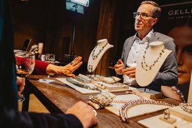 Verkaufsstand mit Schmuck auf Holzplatte, darunter Halsketten, Armbänder und Ringe, im Hintergrund ein Mann in Anzugjacke.