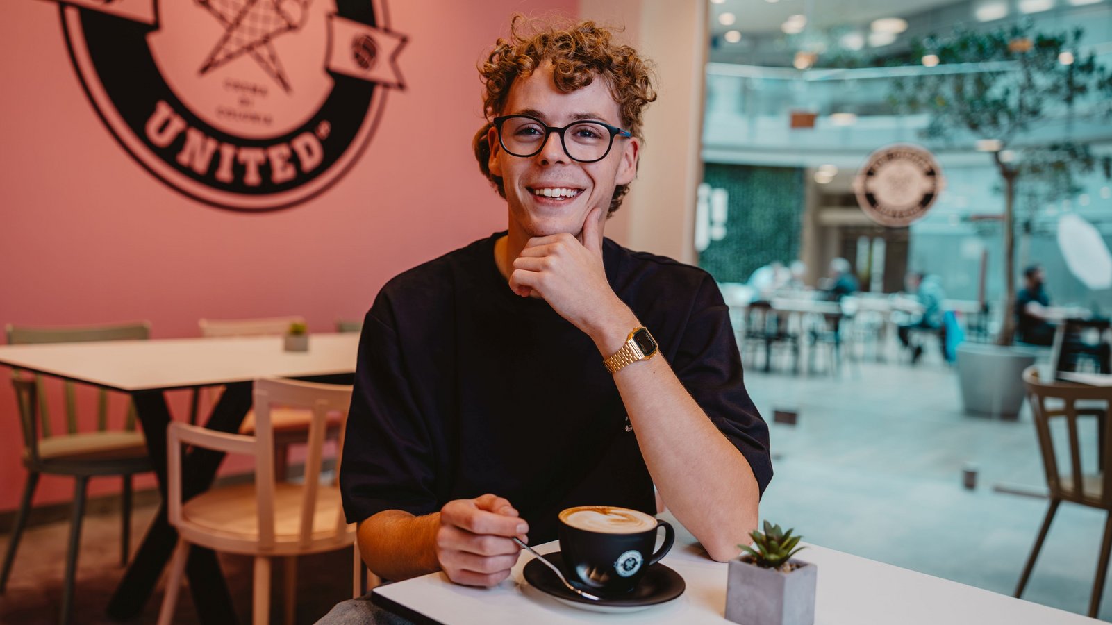 Julian Wöllenstein sitzt an einem Tisch in einem Café, hält eine Tasse Kaffee und trägt eine goldene Uhr am linken Handgelenk.