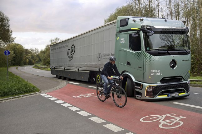 Mercedes-Benz Actros fährt an einem Radweg mit rotem Fahrradschutzstreifen vorbei, auf dem ein Radfahrer mit Helm unterwegs ist