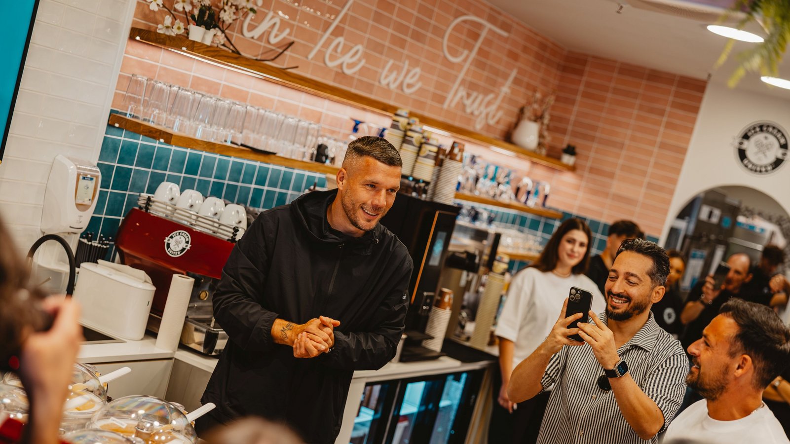Mann in schwarzer Jacke steht hinter Theke mit Eisbehältern, umgeben von mehreren Personen in Eiscafé mit hellen Fliesen und Regalen mit Gläsern.
