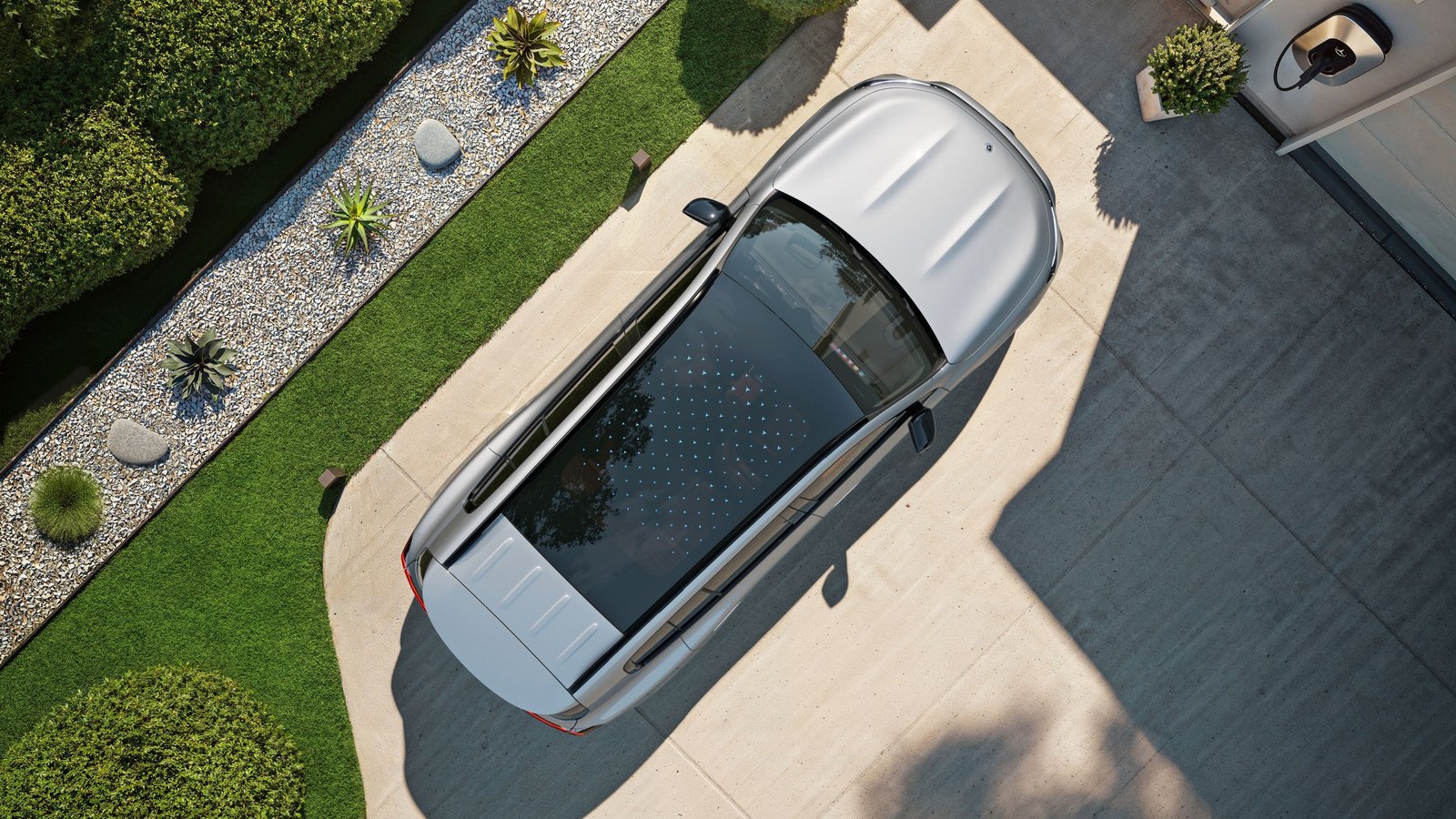 Vogelperspektive auf ein silbernes Auto mit schwarzem Glasdach, das auf einer Einfahrt neben einem Haus steht, umgeben von gepflegtem Grün und Kiesbeeten.