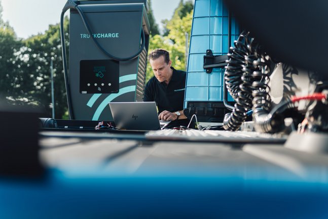 Mann mit Laptop an einer Mercedes-Benz eActros Truckcharge-Ladestation neben einem blauen Lkw-Aufbau