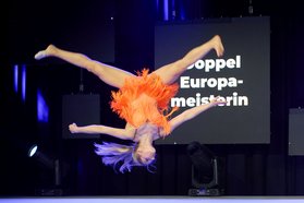 Karina Schönmeier in orangefarbenem Kostüm führt einen gestreckten Salto mit gespreizten Beinen auf Bühne vor schwarzem Hintergrund aus