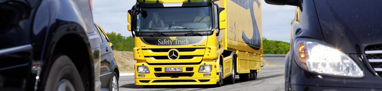 Gelber Mercedes-Benz Lkw mit der Aufschrift 'Safety Truck' fährt auf einer Straße zwischen zwei schwarzen Pkw