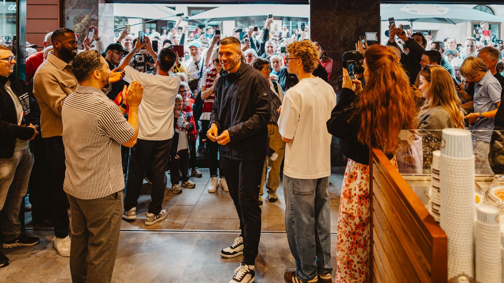 Menschenmenge in einem Geschäftseingang, einige klatschen, andere fotografieren mit Smartphones, helle Innenbeleuchtung