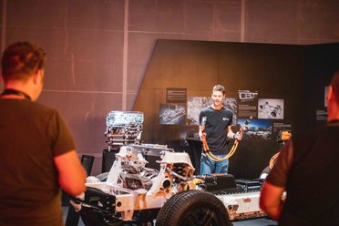 Mann präsentiert vor Publikum das Fahrgestell einer Mercedes-Benz G-Klasse mit sichtbarem Motor und Kabeln in einem Ausstellungsraum.