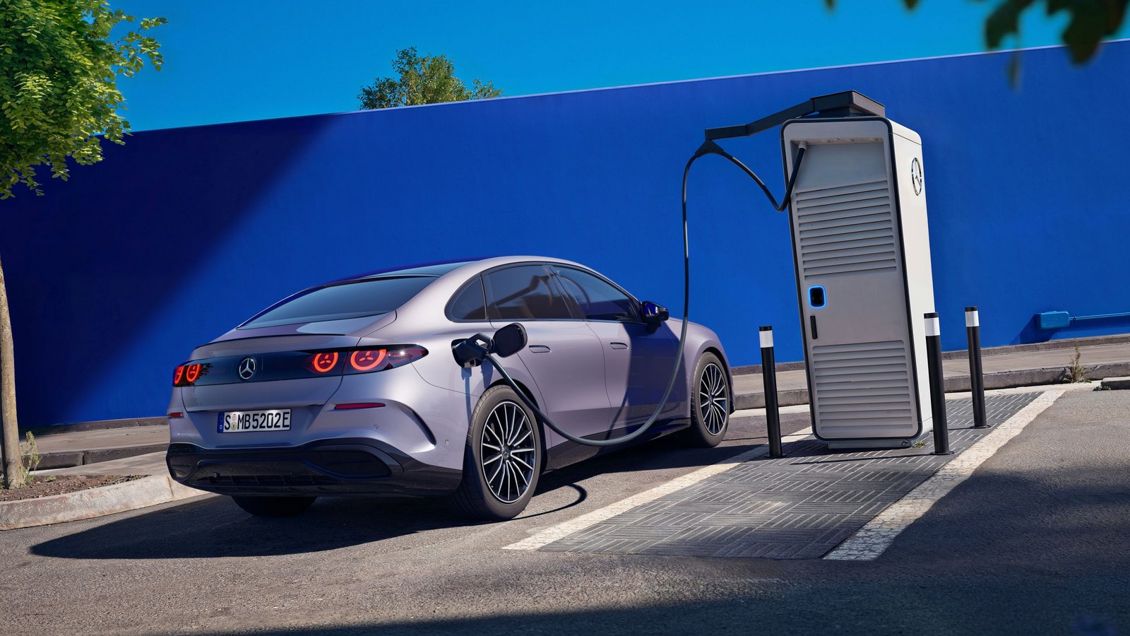 Mercedes-Benz C-Klasse elektrisch lädt an einer Ladestation auf einem Parkplatz vor einer blauen Wand, umgeben von Bäumen und Pflanzen