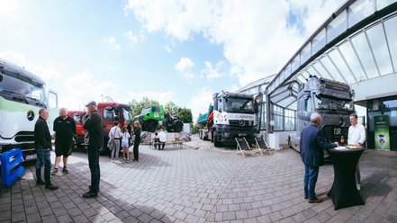Mehrere Personen stehen und unterhalten sich auf einem gepflasterten Freigelände mit mehreren geparkten Mercedes-Benz Trucks und einem Pavillon im Hintergrund.